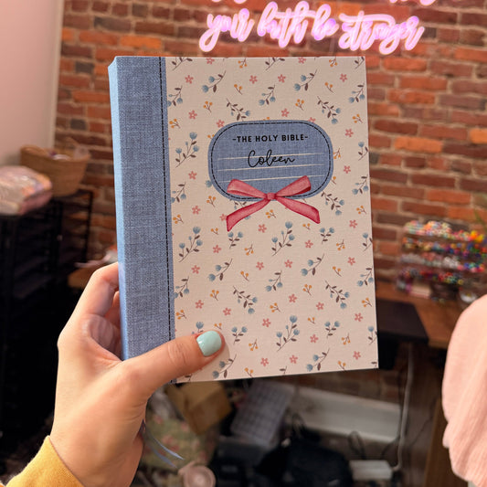 Hand holding a decorative card with floral pattern and blue ribbon against a brick wall background.